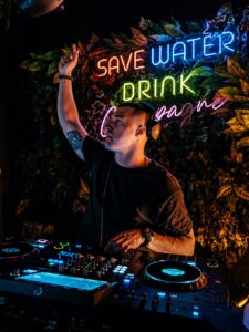 DJ za konsolą pod neonem Save Water Drink Champagne podczas imprezy klubowej